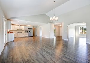 Spacious rambler home interior with vaulted ceiling over glossy laminate floor. Empty light filled dining or living space adjacent to new white kitchen room features pale grey walls. Northwest, USA